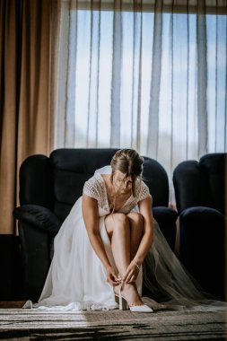 Gelin, beyaz topuklularını giyip, dantelli bir gelinlik ve narin bir bilezik takarak, hafifçe aydınlatılmış bir odanın penceresinde oturur..