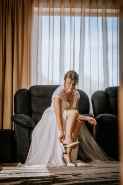 Gelin, beyaz topuklularını giyip, dantelli bir gelinlik ve narin bir bilezik takarak, hafifçe aydınlatılmış bir odanın penceresinde oturur..