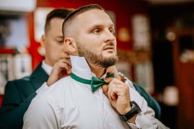 Valmiera, Letonya - 9 Ağustos 2024 - Bir damat yeşil papyonunu bir sağdıcın yardımıyla şık bir iç mekan düzeninde düğün öncesi birlikte hazırlanırken düzeltir..