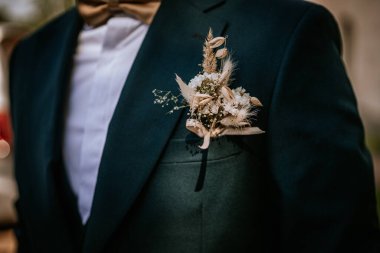 Kurutulmuş çiçeklerden yapılmış zarif bir yaka çiçeğinin yakın çekimi ve kurdeleyle bağlanmış, uygun papyonlu koyu renk bir cekete tutturulmuş.