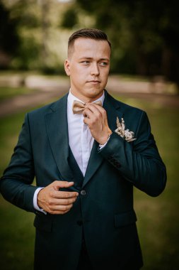 Valmiera, Letonya - 9 Ağustos 2024 - Damat, düğün gününde sakin ve zarif görünen koyu renk bir takım elbise içindeki bir portre için poz verirken altın papyonunu açık havada ayarlar..