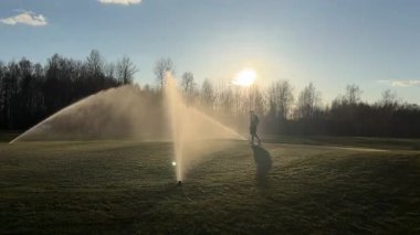 Bir insan gün doğumunda ya da batımında golf sahasında yürür, sisli havada ve uzun gölgeler çiğ çimenler üzerinde uzanır..