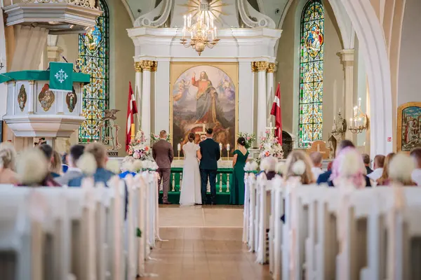 Valmiera, Letonya - 9 Ağustos 2024 - Gelin ve damat mihrapta rahip ve misafirlerle birlikte büyük bir kilisenin içinde beklerken nikah töreni.
