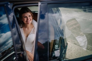 Gülümseyen gelin, düğün gününde arabada oturur. Damat camda görünür bir yansımayla neşeli ve samimi bir anı yakalar.