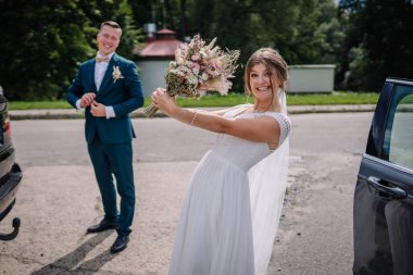 Neşeli gelin buketi ile heyecanla poz verirken gülen damat dışarıda eğlenceli bir düğün anında takım elbisesini arka planda ayarlıyor..