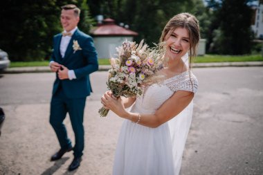 Neşeli gelin kırsal bir buket tutuyor ve arka planda damat ile birlikte açık havada bir düğün anında gülümsüyor..