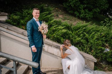 Damat kameraya gülümserken buketi tutar, gelinlik giymiş gelin diz çöker ve taştan bir duvara mavi keçeli kalemle yazar, yeşillik yakınlarında..