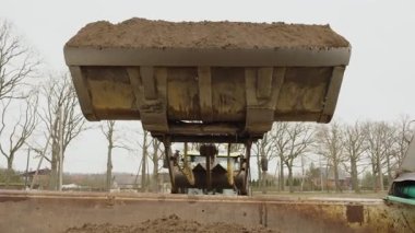 Valmiera, Letonya - 16 Nisan 2025 - Bir cephe yükleyicisi, ağır makineler ve malzeme transferini gösteren bir inşaat sahasındaki bir kamyon yatağına bir kova dolusu ıslak toprak boşaltıyor..