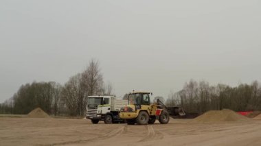 Valmiera, Letonya - 16 Nisan 2025 - Yükleyici ve boşaltma kamyonu doldurma işleminden sonra kısa bir süre durakladı; kamyon yükleyici olarak yola çıktı. Geniş açı, sonra yakın plan. Yumuşak kayma hareketi.