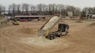 Valmiera, Letonya - 16 Nisan 2025 - Bir çöp kamyonu, arka planda bir tekerlek yükleyicisi çalışırken, büyük bir inşaat sahasında bir yığın kum boşaltır..