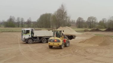Valmiera, Letonya - 16 Nisan 2025 - Sarı bir Volvo yükleyicisi, içinde bir işçiyle birlikte ağaç ve evlerle dolu kırsal bir alanda kum yığınlarıyla çevrili beyaz bir kamyona kum boşaltıyor..