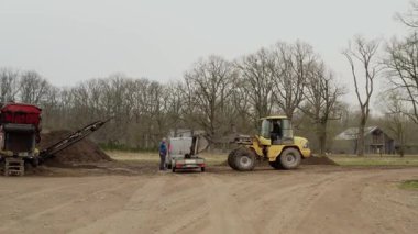 Valmiera, Letonya - 16 Nisan 2025 - Volvo doldurucusuna yaklaşan yavaş kayma sahnesi yakınlarda duran bir adam ve arka planda kırmızı bir ekran ile küçük bir kamyona toprak döküyor.