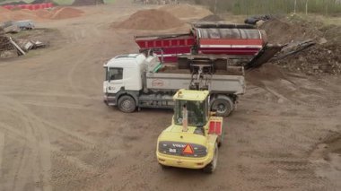 Valmiera, Letonya - 16 Nisan 2025 - Sarı bir Volvo yükleyici, kırsal bir inşaat alanındaki kırmızı toprak tarayıcısının yanındaki beyaz bir çöp kamyonunu görünür lastik izleri ve malzeme yığınlarıyla doldurdu.