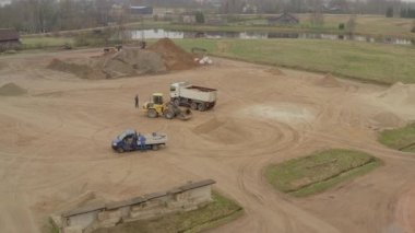 Valmiera, Letonya - 16 Nisan 2025 - Büyük bir kırsal alanda beyaz bir kamyonu kumla dolduran bir Volvo yükleyicisinin yukarıdan aşağıya daha yakın görüntüsüne geniş bir hava geçişi. İki işçi var..