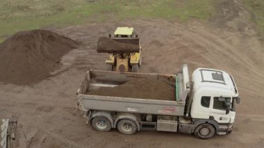 Valmiera, Letonya - 16 Nisan 2025 - Sarı yükleyici kırsal bir bölgedeki beyaz bir çöp kamyonuna toprak yığınları ve lastik izleriyle birlikte büyük bir kova toprak boşaltıyor..