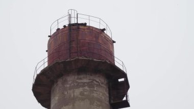 Metal merdiveni ve korkuluğu olan paslı bir endüstriyel su kulesine yakın plan, gri, bulutlu bir gökyüzüne karşı duruyor..