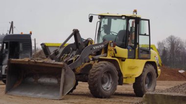 Valmiera, Letonya - 16 Nisan 2025 - Sarı Volvo ön yükleyici bir inşaat sahasına park edilmiş, arka planda kum dolu, kamyonlarla ve malzeme yığınlarıyla dolu kova.