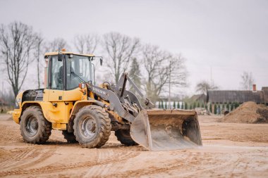 Valmiera, Letonya - 17 Nisan 2025 - Sarı bir Volvo ön-uç yükleyicisi kumlu bir inşaat sahasında duruyor, kova aşağıda, arka planda kırsal binalar ve yapraksız ağaçlar var..