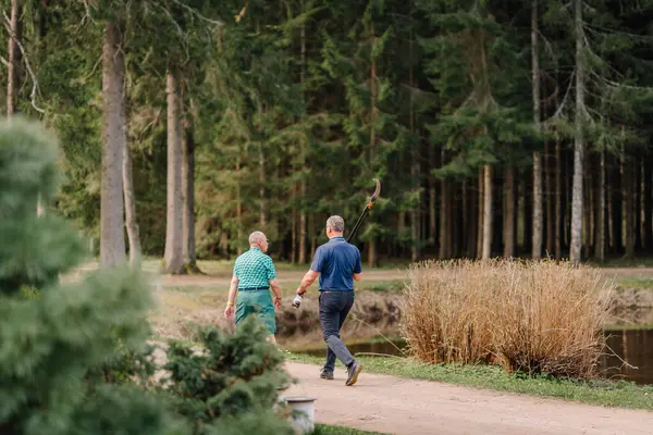 Sigulda, Letonya - 19 Nisan 2025 - Biri elinde bir sırıkla yürüyen iki adam, ağaçlarla çevrili ve huzurlu bir doğal manzara.