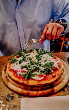 Milano, İtalya - 18 Ağustos 2022 - Mavi gömlekli bir adam, üzerinde roka, prosciutto ve kıyılmış peynir bulunan taze pişmiş bir pizzayı, sıcacık bir restoranda yemeye hazırlanıyor..