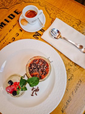 Milan, İtalya - 10 Mart 2025 - Beyaz bir tabakta sunulan böğürtlen ve nane yapraklı zarif tiramisu tatlısı, yanında bir fincan espresso ve ahşap bir masada bir kurabiye.