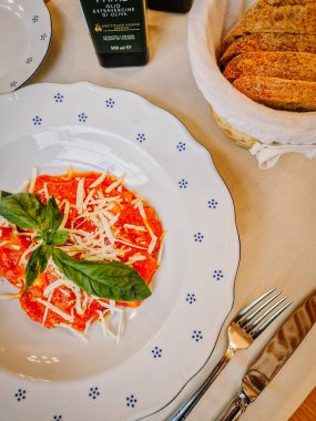 Milan, İtalya - 10 Mart 2025 - Beyaz bir tabakta domates soslu ravioli, rendelenmiş peynir ve fesleğen, sıcak bir yemek ortamında rustik ekmek ve zeytinyağı ile servis edilir..