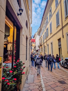 Milan, İtalya - 10 Mart 2025 - Sarı binalar, yayalar, çiçekler ve yakındaki bir restoranın varlığını gösteren tabelalar içeren dar Avrupa caddesi.