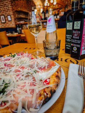 Milan, İtalya - 10 Mart 2025 - Prosciutto ve peynirli gurme pizzaya yakın çekim, sıcak bir restoranda beyaz şarap ve zeytinyağı ile servis edilir.
