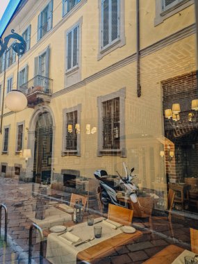 Milan, İtalya - 5 Nisan 2025 - Restoran vitrininde geleneksel sarı bir Avrupa binasının yansıması, sehpaları, sıcak iç mekânları ve park etmiş bir scooter.