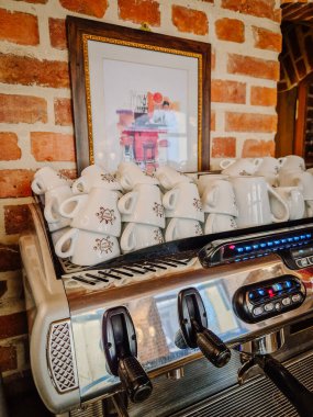 Milan, İtalya - 5 Nisan 2025 - Kırsal bir kafe veya restoranda seramik bardakları istiflenmiş Espresso makinesi, üzeri çerçeveli sanat eserleriyle kaplı tuğla bir duvara karşı