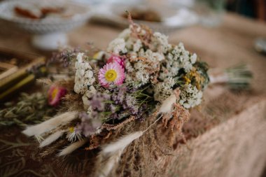 Valmiera, Letonya - 9 Ağustos 2024 - Pembe çilek çiçekleri de dahil olmak üzere kurumuş ve taze çiçekleri desenli bir masa örtüsü üzerinde olan rustik bir gelin buketinin yakın çekimi. Yumuşak, doğal düğün ayrıntıları.