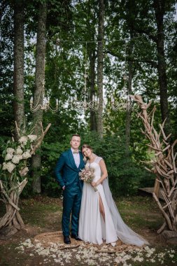 Gelin ve damat ormanda çiçek kemeri ve avizelerle poz veriyorlar. Doğal ve kırsal dekorlu romantik açık hava seremonisi.