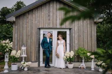 Gelin ve damat çiçekleri ve fenerlerle süslenmiş ahşap bir kulübede gülümsüyor. Büyüleyici bir kırsal düğün anı.