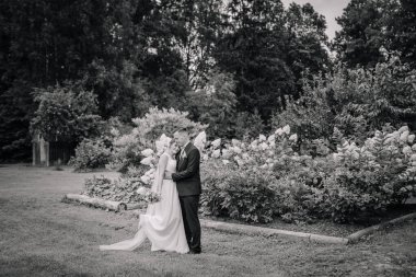 Gelin ve damat, çiçek açan bir bahçede siyah beyaz bir bahçede sessizce kucaklaşırlar. Açık havada romantik ve zamansız bir an..