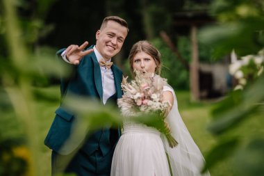 Gelin ve damat yeşilliğin arkasında neşe içinde poz veriyor, gelin öne buket tutuyor ve damat barış işareti gösteriyor. Eğlenceli, açık havada samimi bir düğün..