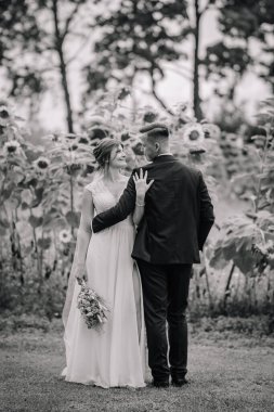 Gelin ve damat, siyah beyaz bir fotoğrafta ayçiçeği tarlasının önünde sevgiyle kucaklaşırlar. Yumuşak ve zamansız kırsal düğün portresi.