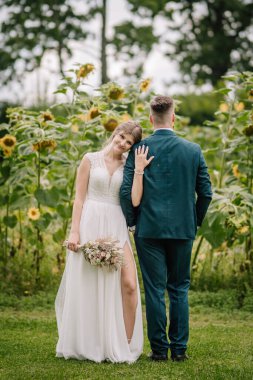 Gelin, bir ayçiçeği tarlasının önünde buketi tutarak damatların sırtına hafifçe yaslanır. Kırsal kesimde neşeli ve hassas bir düğün anı.