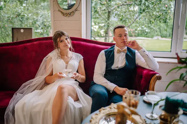 Gelin ve damat, elinde çay fincanlarıyla klasik kırmızı bir kanepede oturup sessiz bir evin tadını çıkarıyorlar. Zarif, rahat ve şık bir düğün ortamı..