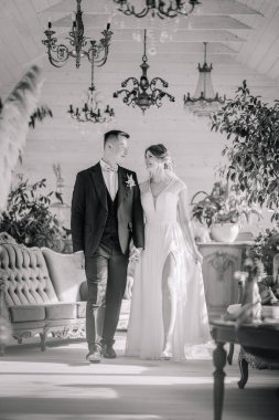 Bir gelin ve damadın zarif siyah beyaz fotoğrafı, el ele yürüyorlar, gülümsüyorlar, avizeler ve yeşilliklerle dolu klasik bir ev ortamında..