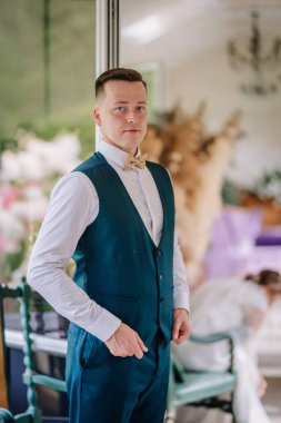 Yeşil yelekli ve papyonlu bir damat törenden önce güvenli bir şekilde içeride poz verir. Gelin arka planda bulanıklaşır ve hazırlanır..
