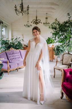 Valmiera, Letonya - 9 Ağustos 2024 - Yüksek yarıklı zarif beyaz elbiseli, gülümseyen bir gelin avize ve klasik mobilyalarla dolu, kapalı, eski bir odada poz veriyor..