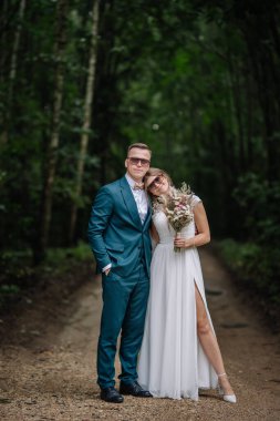 Gelin ve damat, güneş gözlüğü takarak ve sevgiyle gülümseyerek orman yolunda birbirlerine yakın dururlar. Gelin, elinde kır çiçeği buketi tutarken..