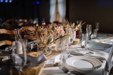 Valmiera, Letonya - 9 Ağustos 2024 - Kurumuş çiçek süslemeleri, beyaz tabaklar, cam eşyalar ve resmi bir etkinlik için yumuşak doğal ışık içeren zarif bir köy masasının yakın çekimi.