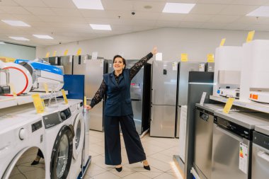 Valmiera, Letonya - 23 Nisan 2025 - Çamaşır makineleri, buzdolabı, bulaşık makineleri ve ev aletleriyle çevrili bir elektronik mağazasında yükseltilmiş koluyla poz veren neşeli kadın.