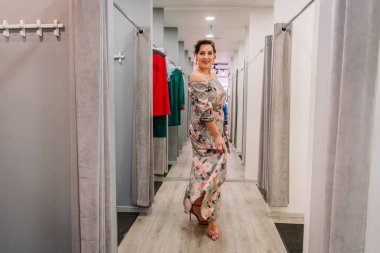 Valmiera, Letonya - 23 Nisan 2025 - Butik soyunma odasının koridorunda çiçekli bir maksi elbiseyle yürüyen gülen kadın, etek kenarını kendine güvenen bir zarafetle tutuyor..