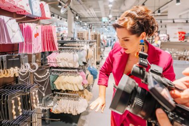 Valmiera, Letonya - 23 Nisan 2025 - Pembe ceketli kadın bir moda mağazasında aksesuar arkaplanda görünür mücevherler ve şapkalar sergilenirken filme alınıyor.