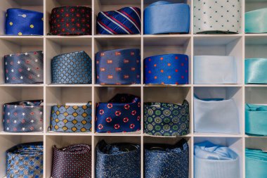 Beyaz bir rafta desenli, koyu mavi, siyah ve açık turkuaz erkek kravatları sergilenmekte, şık bir kravat koleksiyonu sergilenmektedir.