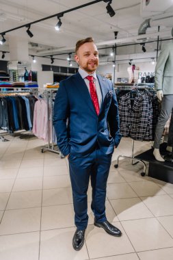 Valmiera, Letonya - 23 Nisan 2025 - Kırmızı kravatlı ve cep mendili olan lacivert takım elbiseli bir adam modern bir erkek giyim mağazasında poz veriyor..