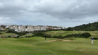 Malaga, İspanya - 24 Ocak 2025 - Üç golfçü bulutlu gökyüzünün altında arka planda manzaralı bir yamaç kasabasıyla yeşil bir golf sahasına atış yapmaya hazırlanıyor.