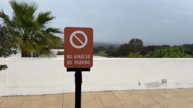 Yağmurlu ve bulutlu bir günde, arka planda palmiye ağacı ve golf sahası olan beyaz bir duvarın yanında İspanyolca ve İngilizce park işareti yok..